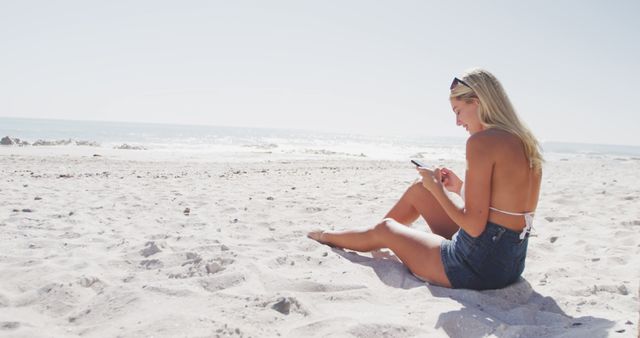 Blonde woman sitting on a sandy beach on a sunny day, looking at her smartphone. She is wearing a bikini top and denim shorts. The image captures a serene and relaxing moment, perfect for illustrating concepts such as leisure, summer vacations, technology use in nature, and solo enjoyment time. Ideal for travel websites, summertime promotions, technology in nature contexts, and lifestyle blogs.