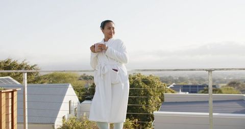 Asian woman enjoying morning coffee on rooftop terrace in white bathrobe