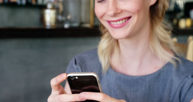 Close-up of smiling woman using smartphone indoors. She is holding device with both hands, engaging in a conversation. Great for illustrating themes of modern technology, communication, and everyday casual moments. Useful for articles on mobile technology, social media interaction, or lifestyle blogs.