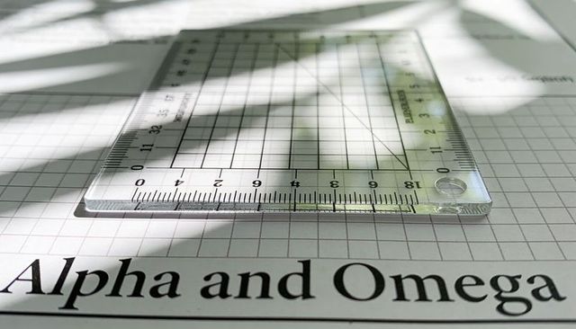 Clear acrylic square ruler resting on grid paper showing scales and alpha and omega