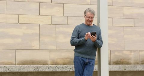 Mature man leaning on post checking smartphone against modern beige facade outdoors