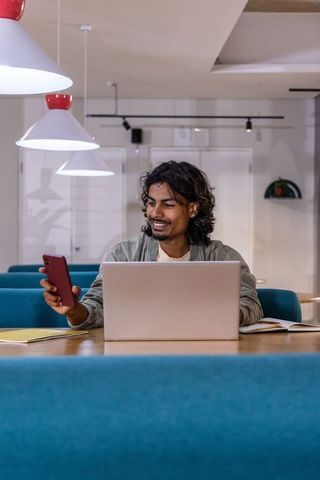 Indian man smiling, checking red phone while typing on laptop in coworking lounge