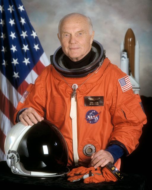 A senior astronaut wearing an orange NASA spacesuit stands in front of an American flag and a space shuttle model while holding his helmet. Ideal for use in articles and educational materials about space exploration, NASA missions, or senior figures in space history.