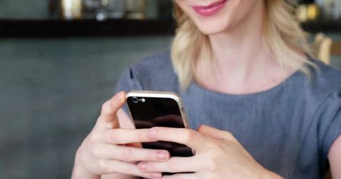 Smiling woman holding smartphone tapping screen while sitting in cozy cafe lounge with ambient light