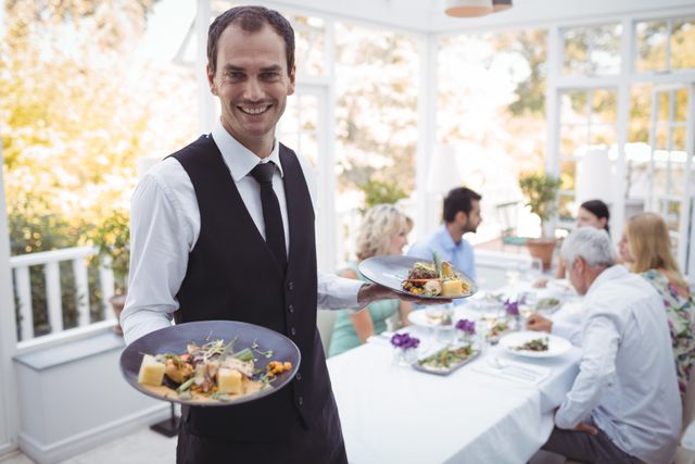 Waiter in formal attire smiling while holding a food tray in an elegant restaurant. Ideal for use in hospitality industry promotions, restaurant advertisements, customer service training materials, and dining experience brochures.