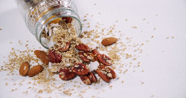 A variety of nuts and seeds are spilled from a glass jar onto a white surface, showcasing a mix of almonds, pecans, and grains. This arrangement emphasizes the concept of healthy eating and the importance of including nuts and seeds in a balanced diet.