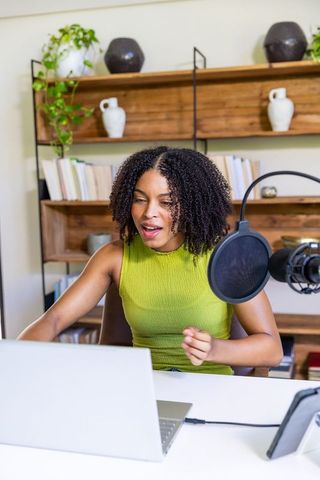 African american woman recording podcast in bright home studio with microphone and laptop