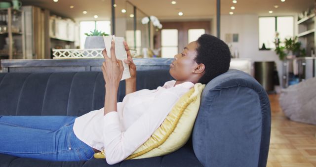 This image shows an African American woman lying on a couch and reading a book in a modern living room. It could be used in lifestyle blogs, articles about leisure activities, home decor magazines, or relaxation-focused advertisements and social media posts.