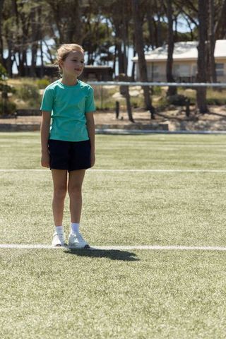 Young schoolgirl standing on sports turf sideline wearing turquoise t-shirt and navy shorts