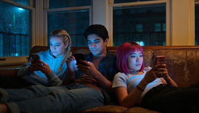 Late-night friends lounging on leather sofa scrolling phones under rainy window glow