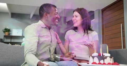 Couple sharing cake on sofa celebrating birthday with candles, laughter and cozy romance