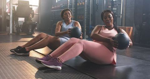 Diverse women twisting with medicine balls in gym, partner core training for strength