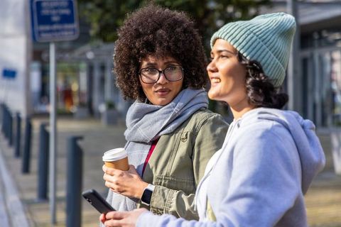 Sunlit urban friends chatting while holding coffee and smartphone multicultural lifestyle