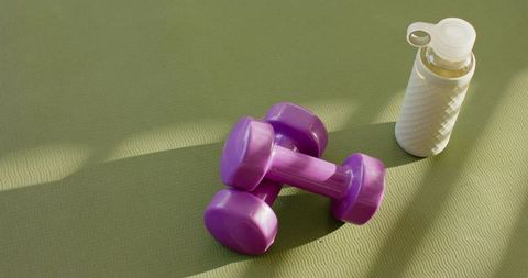 Purple dumbbells on green yoga mat with white water bottle casting warm shadows