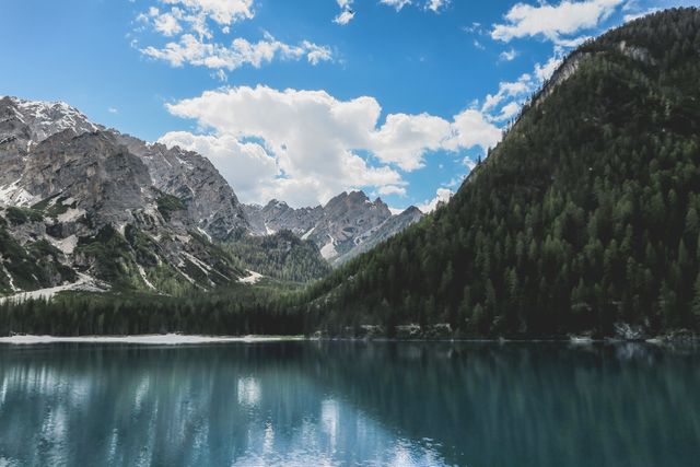This image captures a tranquil mountain lake with a perfect mirror reflection of the surrounding forest-covered mountains and a bright, cloud-filled sky. Ideal for travel blogs, nature documentaries, tourism advertisements, and relaxation or mindfulness content showcasing the beauty and serenity of natural landscapes.