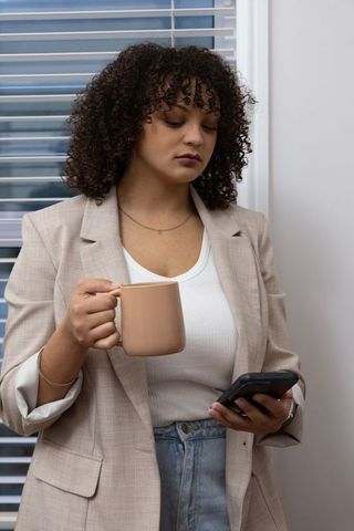 African american professional holding mug and checking smartphone by office blinds