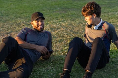 Two men relaxing on grass sharing phone and headphones after workout at golden hour