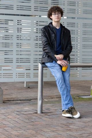 Teenager leaning on railing holding orange juice and snack wearing leather jacket, jeans
