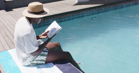 Man reading paperback poolside wearing straw hat and sunglasses for summer relaxation