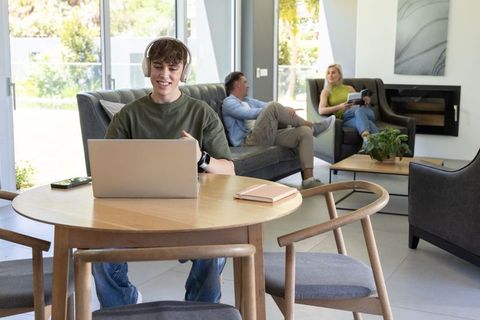 Teenager using laptop and headphones at round table while parents relaxing in modern living room