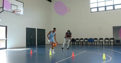 Male basketball player dribbling through cones while coach instructing on indoor court