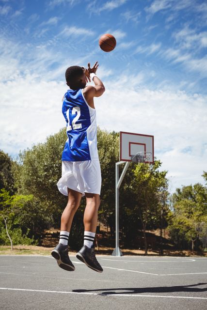 Teenage basketball player making a jump shot on an outdoor court during a sunny day. Ideal for use in sports-related content, youth athletic programs, fitness campaigns, and motivational materials.