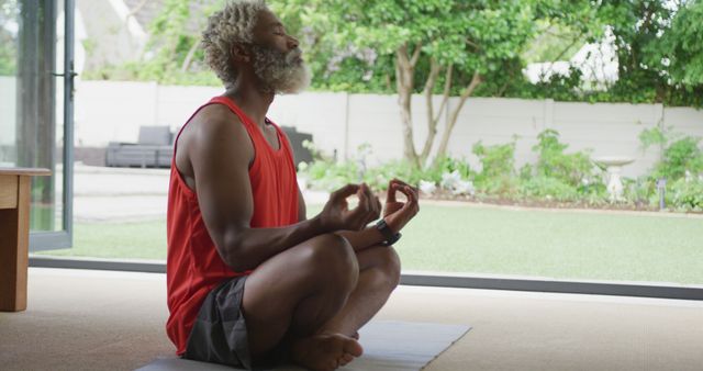 Senior man meditating in a seated yoga pose at home with a view of a garden. Promotes relaxation, mindfulness, and a healthy lifestyle. Ideal for content related to wellness, aging gracefully, indoor-outdoor spaces, and healthy habits.