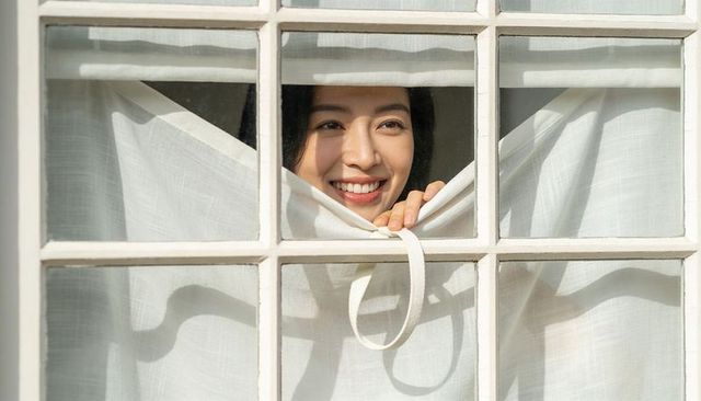 Asian woman peeking through linen curtain, smiling with sunlight on face at window