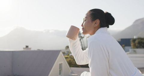 Mid-adult asian woman sipping from pink mug on balcony wearing white robe at mountain sunrise glow