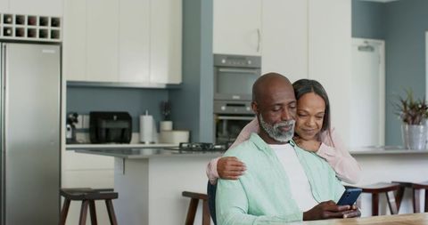 Mature couple cuddling while using smartphone in bright modern home kitchen