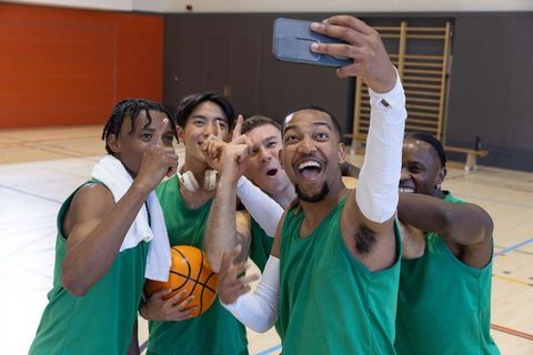 Diverse basketball teammates celebrating victory and capturing selfie on indoor court showing teamwo
