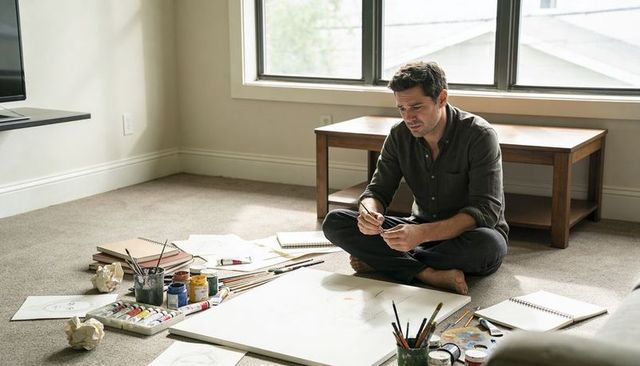 Male artist sitting cross-legged working on canvas with paints and sketches