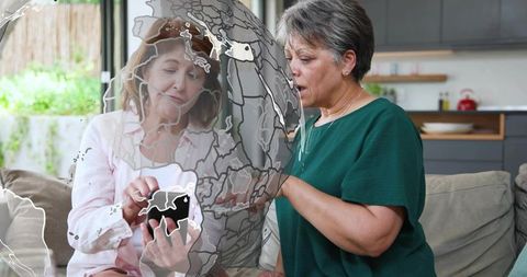 Senior mother and adult daughter sharing smartphone on sofa with globe map overlay