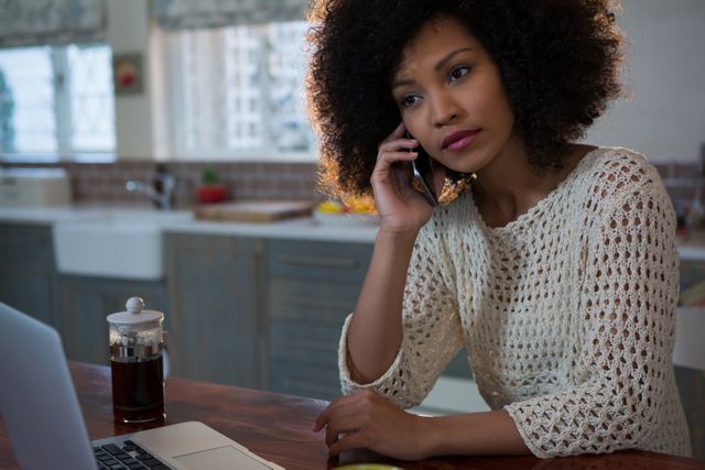 Woman with curly hair talking on mobile phone while sitting at home desk with laptop and coffee. Ideal for illustrating remote work, communication, home office, or casual lifestyle themes.