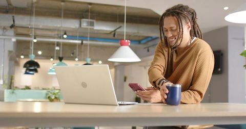 African American man leaning on communal desk, smiling while using smartphone and laptop