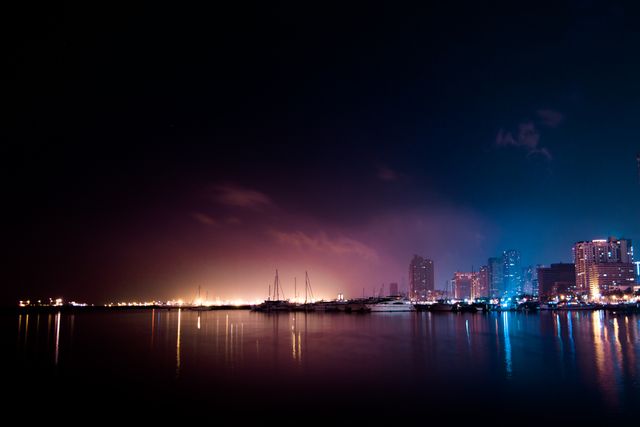 Nighttime view of a city skyline with skyscrapers and buildings reflecting in the calm waters of a river. The harbor features boats and yachts adding a touch of elegance. Ideal for travel websites, urban-themed presentations, background images for advertisements, and articles about city nightlife.