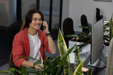 Smiling asian man talking on smartphone while working at modern plant-filled office desk