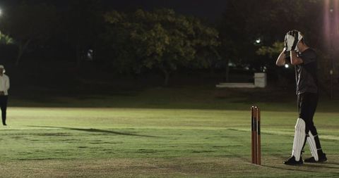 Night cricket batter holding helmet by wooden stumps under floodlights on grass field