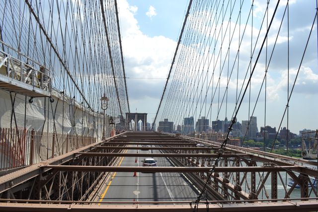 This image shows the perspective view of the Brooklyn Bridge, focusing on its intricate steel cables, pedestrian walkway, and lanes for vehicle traffic. The skyline of New York City is visible in the background underneath a partly cloudy sky. Use this for content relating to urban architecture, travel guides to New York, or articles about iconic landmarks.
