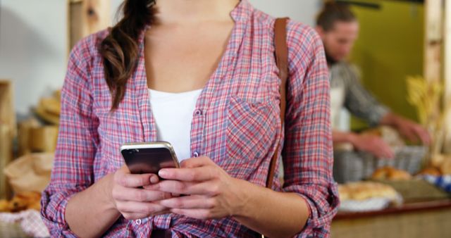 Young woman wearing casual plaid shirt and using smartphone in a market environment. The woman is standing and typing on her phone, highlighting the modern lifestyle and widespread technology use. Ideal for depicting communication, everyday technology, casual fashion, or marketplace imagery. Can be used in advertisements for phone services, lifestyle blogs, or technology-related content.