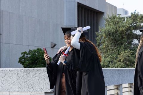 Female graduates celebrating on campus terrace holding diplomas and smartphone