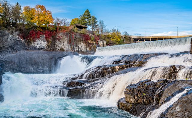 Beautiful waterfall in rural landscape surrounded by vibrant autumn foliage and clear blue sky. Flowing water over rocky falls provides peaceful and scenic view, ideal for travel advertising, nature blogs, and relaxation-themed content.