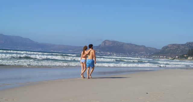 A young Caucasian couple enjoys a romantic walk along a sandy beach, with copy space. Their closeness and the serene coastal landscape create a peaceful and intimate atmosphere.