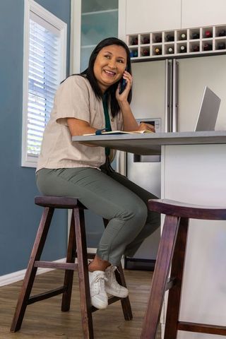 Asian mature woman working from home talking on smartphone at modern kitchen island