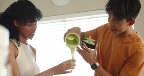 African american woman and asian man pouring green smoothie in sunlit kitchen