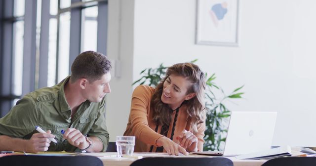 Young male and female coworkers discussing a project in an office setting. There is a laptop on the desk, along with papers and a glass of water. Ideal for illustrating concepts related to teamwork, collaboration, office work, project planning, and professional environments.