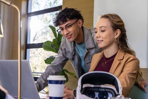 Young diverse coworkers collaborating on laptop with VR headset and coffee in modern office