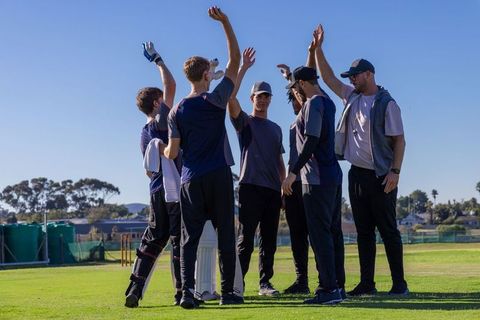 Cricket team huddling and raising hands in celebration on sunlit grass field with coach