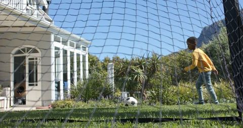 Boy playing soccer in backyard goal net near porch and garden on sunny day