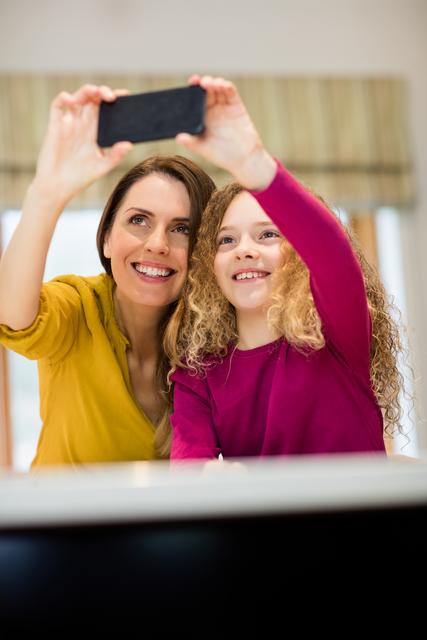 Mother and daughter capturing a happy moment together with a mobile phone. Perfect for use in family-oriented content, parenting blogs, advertisements for mobile phones or technology, and lifestyle articles focusing on family bonding and home life.